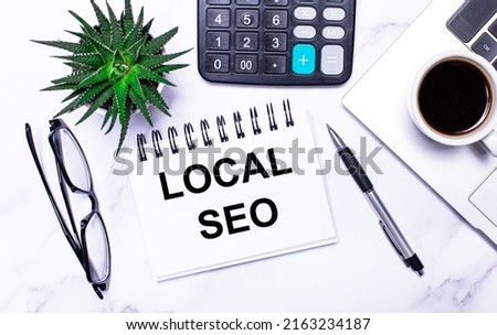 A laptop, glasses, a pen, a calculator, a cup of coffee, a green plant and a white notepad with the text LOCAL SEO lie on a table with task lighting. View from above. Business concept.