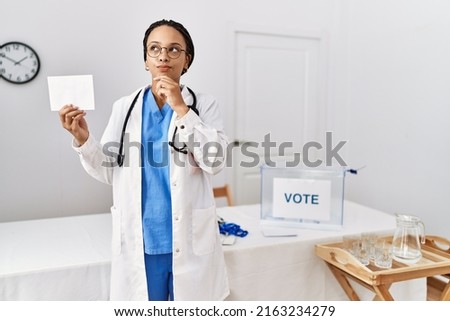 Young african american doctor woman voting holding envelope serious face thinking about question with hand on chin, thoughtful about confusing idea 