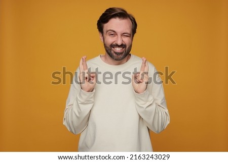 indoor portrait of handsome bearded male posing over orange background keeps his finger crossed, wink, with smile on his face