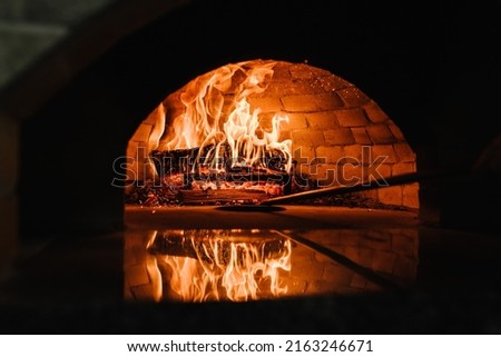 A traditional oven for cooking and baking pizza with a shovel. Firewood burning in the oven. Wood-fired oven. Image of a brick pizza oven with fire. Royalty-Free Stock Photo #2163246671