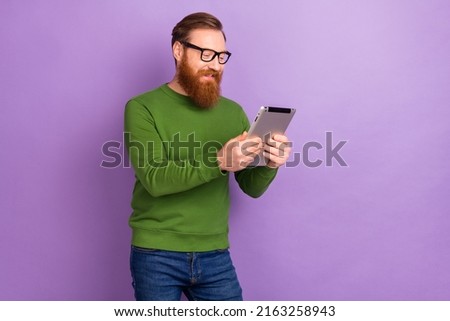 Photo of intelligent focused young male in spectacles searching information in tablet isolated on purple color background