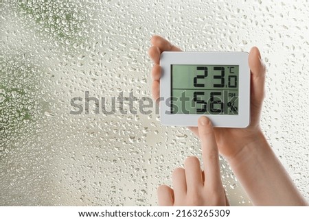 Woman holding digital hygrometer with thermometer near window on rainy day, closeup. Space for text Royalty-Free Stock Photo #2163265309