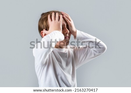 Little boy having a headache. Despair, tragedy. Headache child. Suffering migraine. Headache because stress. Portrait of a sad boy holding his head with his hand, isolated on the gray background. Royalty-Free Stock Photo #2163270417
