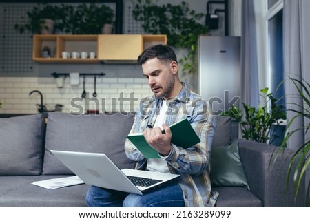 Distance learning online. Young man, student. He studies at home with a laptop, books. Sitting on the couch writes in a notebook online lecture. Listens and watches lessons
