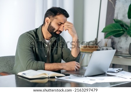Tired exhausted arabic or indian man, office worker, manager or freelancer, sitting at his desk, tired of working in a laptop, overworked, having a headache, closed his eyes, needs rest and break Royalty-Free Stock Photo #2163297479