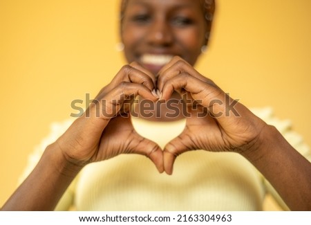 Young woman making a heart shaped symbol while smilling with a yellow background