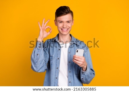 Photo of young cheerful man use mobile show fingers okey symbol promo isolated over yellow color background
