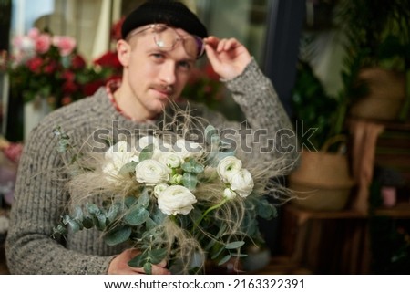 Smiling bearded man with bouquet of white roses in flower shop. Good looking male florist wearing sweater, beanie and eyeglasses holding flower bouquet. Small business concept. High quality image