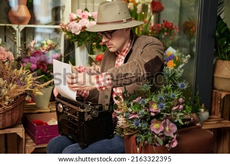 Poet or writer working concept. Cute male person in eyeglasses and hat using old typewriter in flower shop. Suitcase with flower bouquet standing near. High quality image