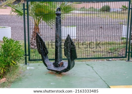 Old black anchor stands vertically close to metal green fence