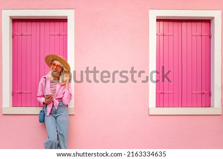 Happy smiling fashionable woman wearing stylish summer outfit holding, using her smartphone, posing near pink color house. Copy, empty space for text

 Royalty-Free Stock Photo #2163334635