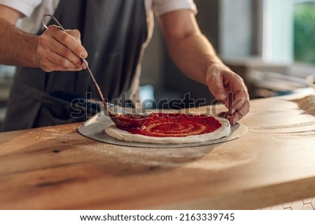 Man worker in a pizza place placing ingredients on pizza base. Special sauce is applied on the pizza dough and pizza ingredients are placed on it.