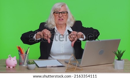 Upset senior mature business woman showing thumbs down sign gesture, expressing discontent, disapproval, dissatisfied, dislike at green chroma key office, using laptop computer. Elderly old freelancer