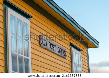 A wooden sign with black letters spelling old post office is affixed to the exterior of a yellow building with green trim. There are vintage glass windows with white trim on the corner of the building