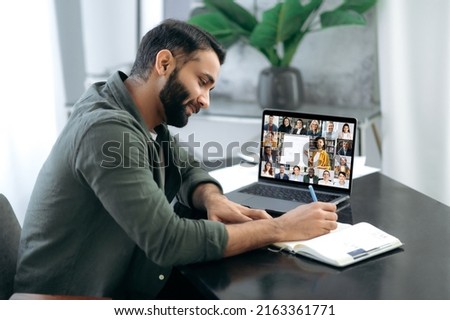 Side view of a successful smart guy listening to an online lecture, taking notes in a notebook, on a laptop screen, a teacher and a group of multiracial people. Online training, webinar Royalty-Free Stock Photo #2163361771