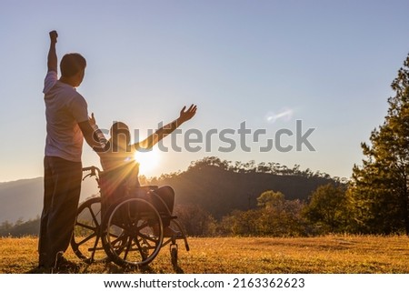 Silhouette of joyful couple in wheelchair raised hands at sunset Royalty-Free Stock Photo #2163362623