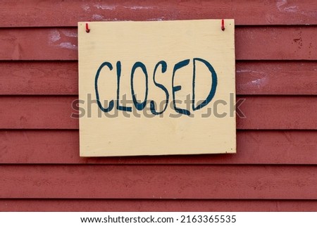 A yellow wooden closed sign with black painted letters hangs on a red wooden building. The narrow red cape cod siding wall is made of painted pine horizontal boards. The word closed is handwritten.  