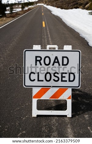 Road closed sign (vertical) on seasonal paved road with snow in the background 