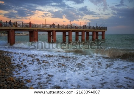 Evening walk along the old high pier over the sea in Adler (Russia) in summer. Fascinating picture of the sunset over the stormy sea. Waves crash against the pebbly shore, forming splashes and foam Royalty-Free Stock Photo #2163394007