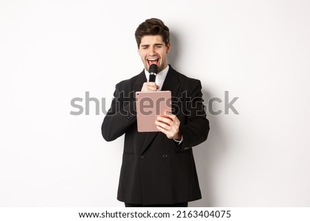 Image of handsome man in black suit, singing karaoke on digital tablet, holding microphone, standing over white background