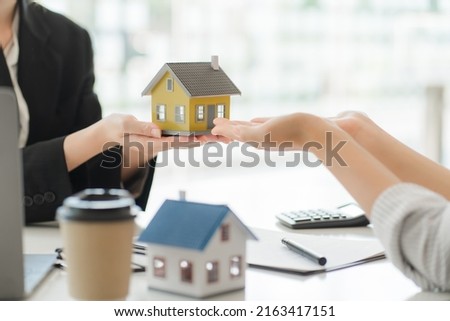 A saleswoman checks the lease and holds a small gray model of house and house keys in preparation for meeting customers to sign the lease. Real estate leasing concept.