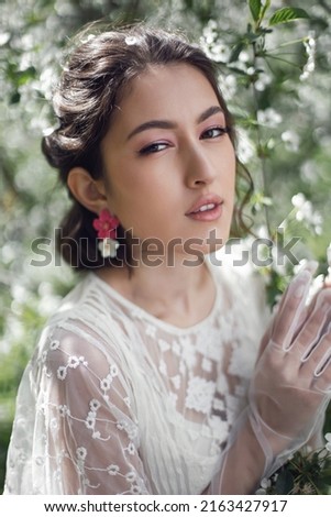 portrait of a young beautiful woman in white clothes standing next to a blooming cherry tree in spring