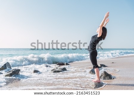 Side view of female stretching body and doing backbend while standing on rock in Tadasana. She is with raised arms on sea coast against blue sky during yoga session Royalty-Free Stock Photo #2163451523