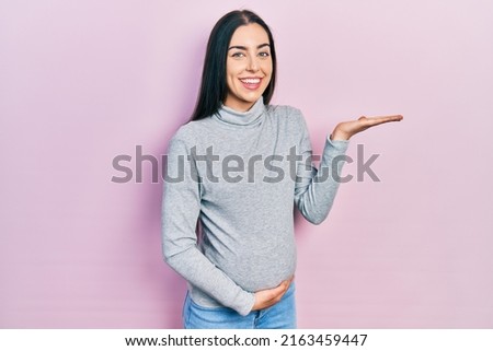 Beautiful woman with blue eyes expecting a baby, touching pregnant belly smiling cheerful presenting and pointing with palm of hand looking at the camera. 