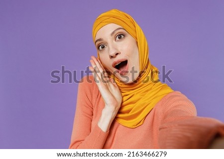 Close up shocked surprised amazed young arabian asian muslim woman in abaya hijab yellow clothes doing selfie shot pov on mobile phone isolated on plain pastel light violet background studio portrait