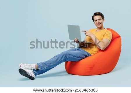 Full body side view fun young man wear yellow t-shirt sit in bag chair hold use work point finger on laptop pc computer isolated on plain pastel light blue background studio. People lifestyle concept