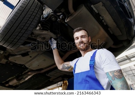 Lower fun strong young male professional technician mechanic man wearing denim blue overalls white t-shirt stand near car lift check technical condition work in vehicle repair shop workshop indoors Royalty-Free Stock Photo #2163466343