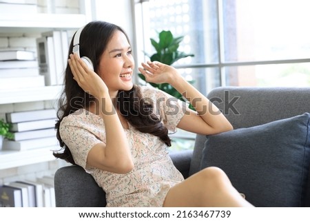 Young happy asian woman wearing headphones relaxing, listening to the music happily from tablet at living room.