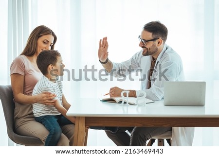 Side view smiling friendly male pediatrician in glasses and white medical coat giving high five to happy little kid boy patient, visiting private clinic with caring young mother, children healthcare.