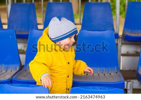 Little boy in yellow jacket playing at empty blue seats in a row at the sport stadium. Toddler child smiling outdoors.