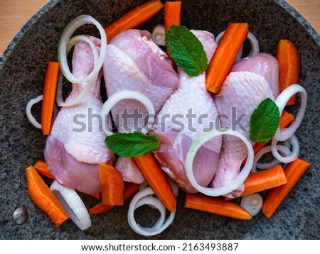Raw chicken drumsticks with carrots, onions and green herbs in frying pan top view. Fresh meat vegetables salad spices cooking. Homemade healthy yummy food ingredient background copy space flat lay.