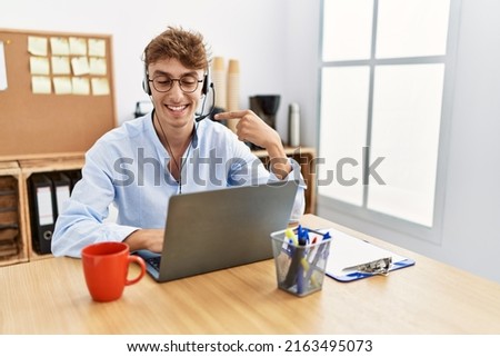 Young caucasian man wearing call center agent headset at the office smiling happy pointing with hand and finger 