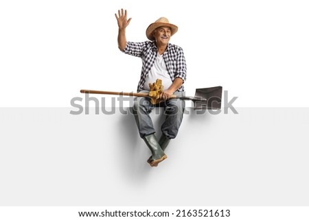 Full length portrait of a mature farmer with a spade sitting on a blank panel and waving isolated on white background Royalty-Free Stock Photo #2163521613