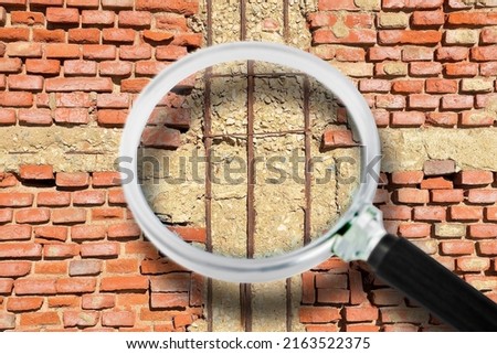 Old reinforced and dangerous concrete structure with damaged and rusty metallic reinforcement bars against a weathered and damaged brick wall - concept image with magnifying glass.