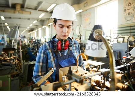 Serious busy skilled lady employee of industrial factory examining equipment while working on lathe machine
