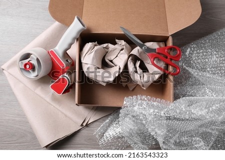 Open box with wrapped items, adhesive tape, scissors, paper and bubble wrap on wooden table, flat lay Royalty-Free Stock Photo #2163543323