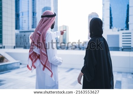 Back view of Arab family or business work colleagues pointing towards city skyline wearing traditional abaya and dress. Muslim Saudi or Emirati couple or team working together Royalty-Free Stock Photo #2163553627
