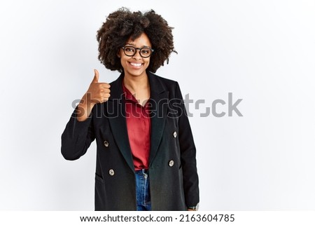African american woman with afro hair wearing business jacket and glasses doing happy thumbs up gesture with hand. approving expression looking at the camera showing success. 