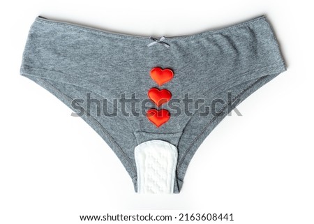 Gray women's panties and white cotton daily sanitary pad and red hearts as mestrual drops on a white background, top view, minimalism. Menstrual cycle. Women's health concept.