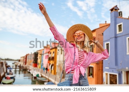 Happy smiling traveler woman posing in street, among colorful houses on Burano island. Girl wearing trendy summer outfit with orange sunglasses, straw hat, pink shirt. Copy, empty space for text Royalty-Free Stock Photo #2163615809