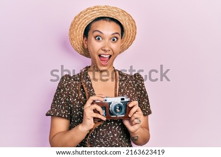 Beautiful hispanic woman with short hair wearing summer hat holding vintage camera celebrating crazy and amazed for success with open eyes screaming excited. 