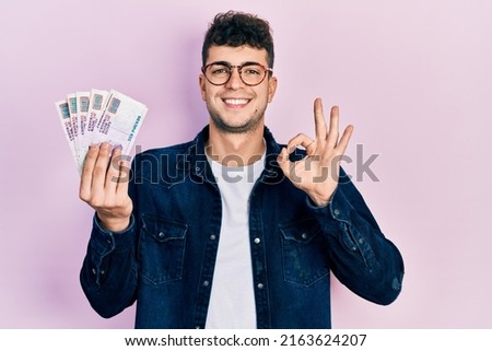 Young hispanic man holding egyptian pounds banknotes doing ok sign with fingers, smiling friendly gesturing excellent symbol 