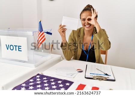 Beautiful hispanic woman holding voting envelop in ballot box smiling happy doing ok sign with hand on eye looking through fingers 