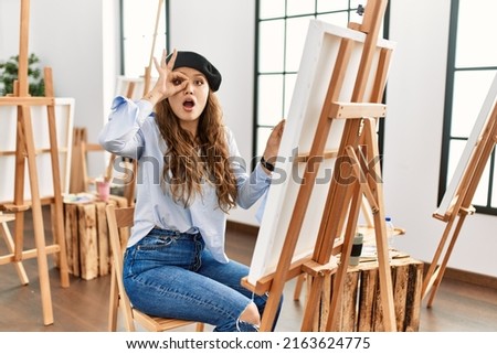 Young hispanic artist woman painting on canvas at art studio doing ok gesture shocked with surprised face, eye looking through fingers. unbelieving expression. 