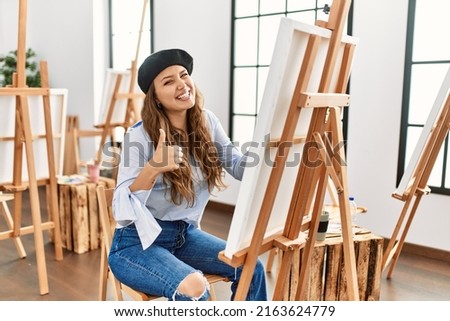 Young hispanic artist woman painting on canvas at art studio approving doing positive gesture with hand, thumbs up smiling and happy for success. winner gesture. 