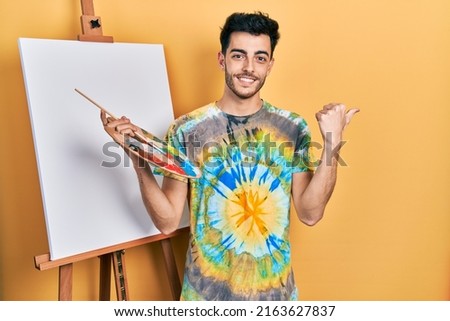 Young hispanic man standing drawing with palette by painter easel stand cheerful with a smile on face pointing with hand and finger up to the side with happy and natural expression 